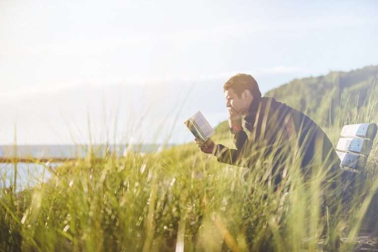 man sitting on bench take a a hard read at a book below his leg is green grass